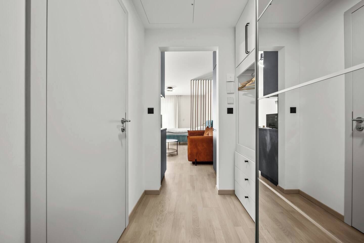 A narrow hallway with a wooden floor leading to a room with a bed, a chair, and a table.