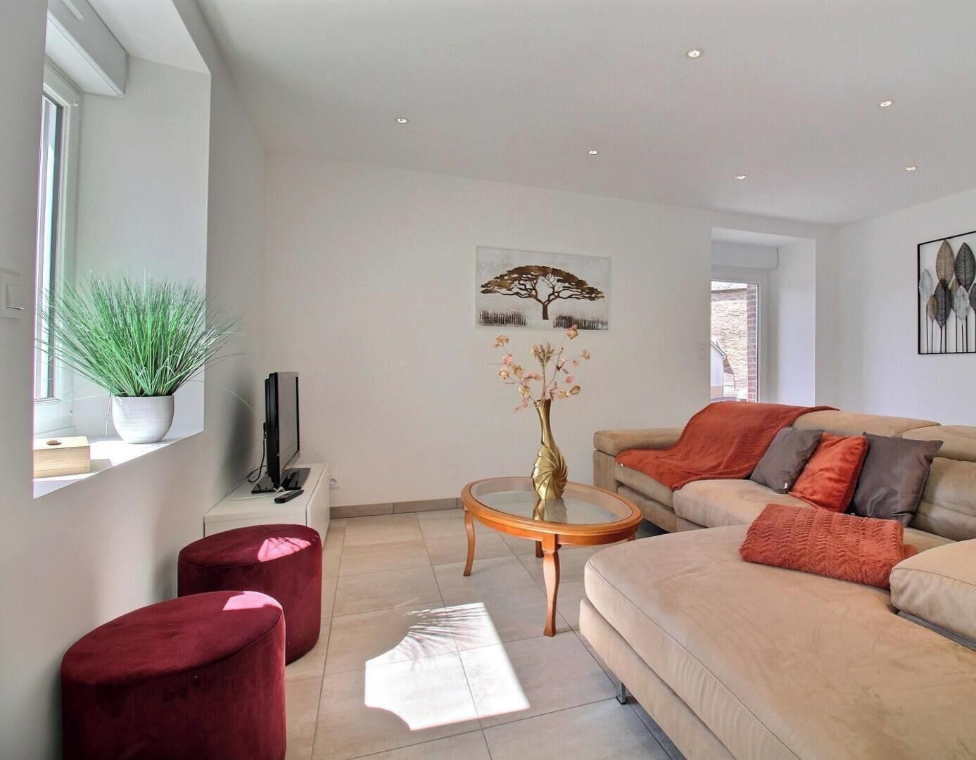 Un salon moderne avec un canapé, une table basse et un téléviseur. Une fenêtre laisse entrer la lumière, et une plante est posée sur le rebord.