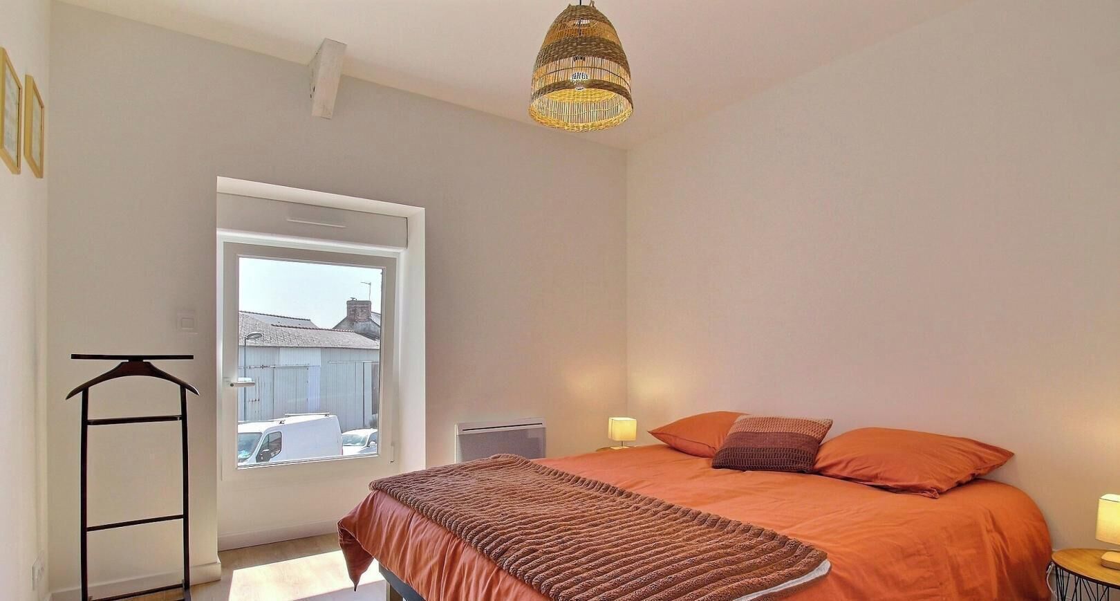 A bedroom with a bed, a hanging light fixture, a radiator, and a window with a view of buildings outside.