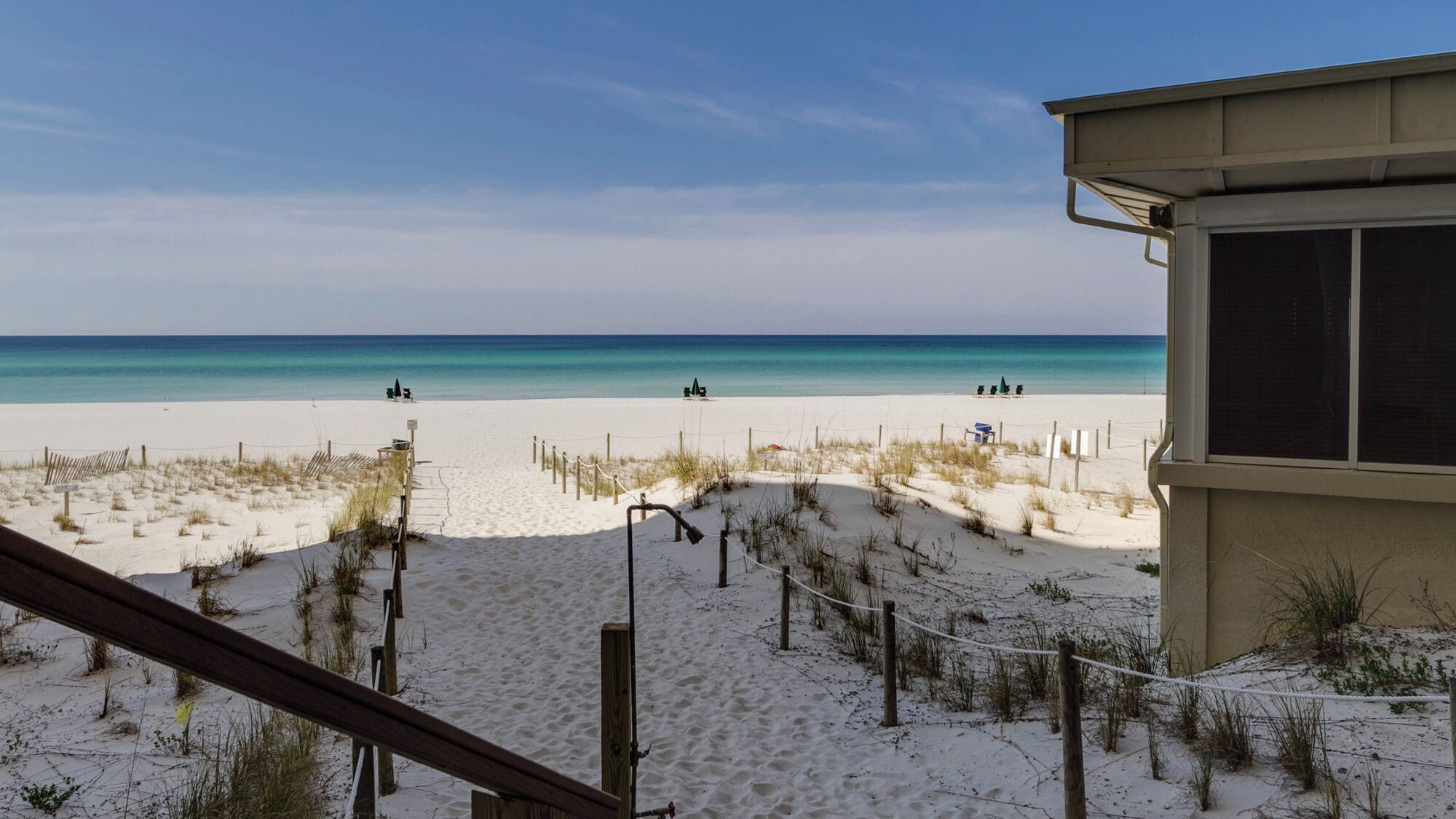 A beach with a sandy path leading to the water, a wooden fence, and a building with a window.