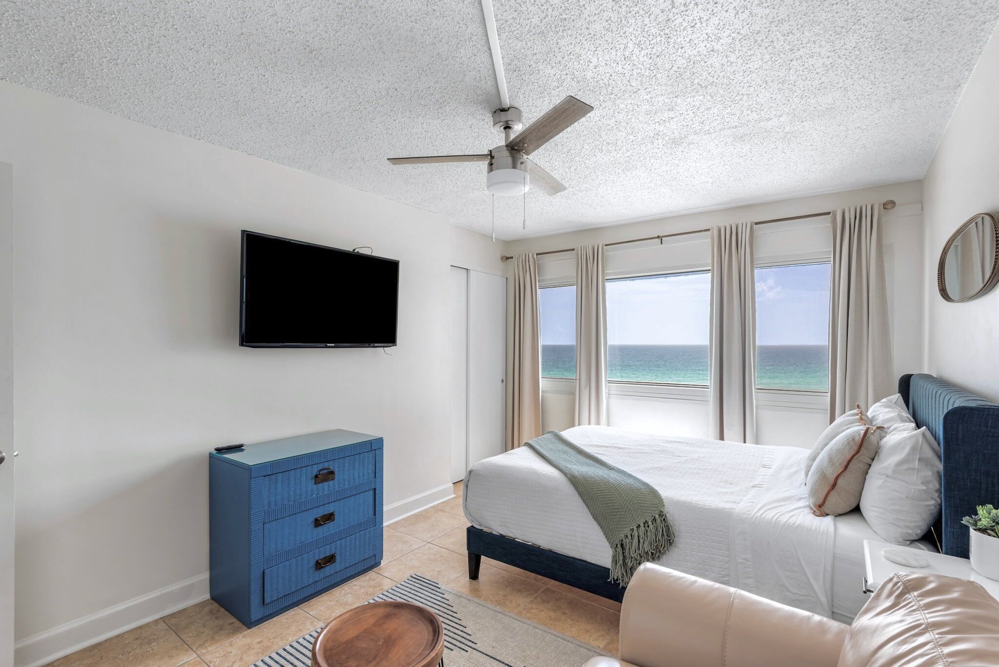 Un dormitorio con cama, televisor, ventilador de techo, cómoda y vista al océano.