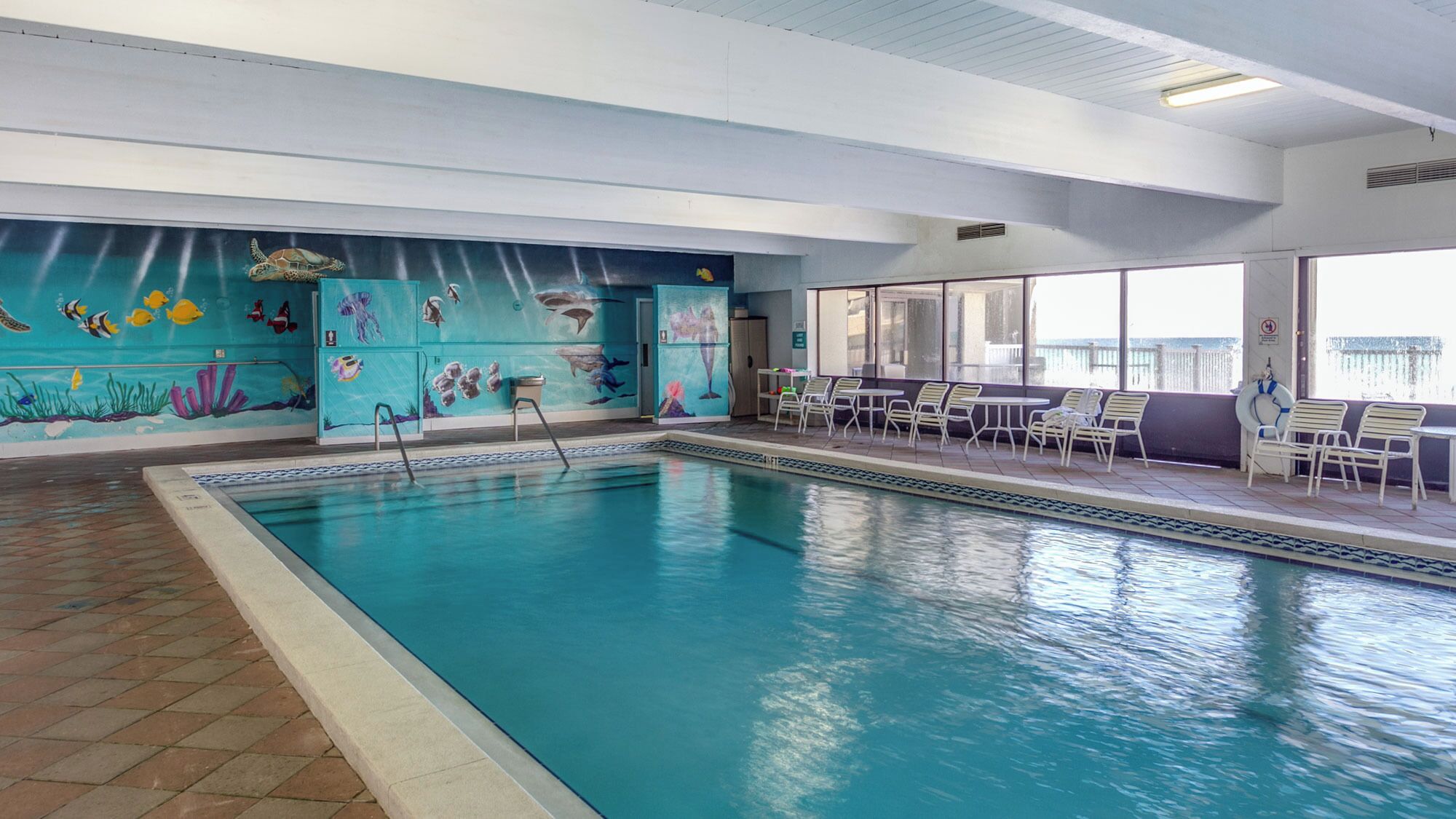 An indoor swimming pool with a mural of marine life on the walls, lounge chairs, and a view of the outside.