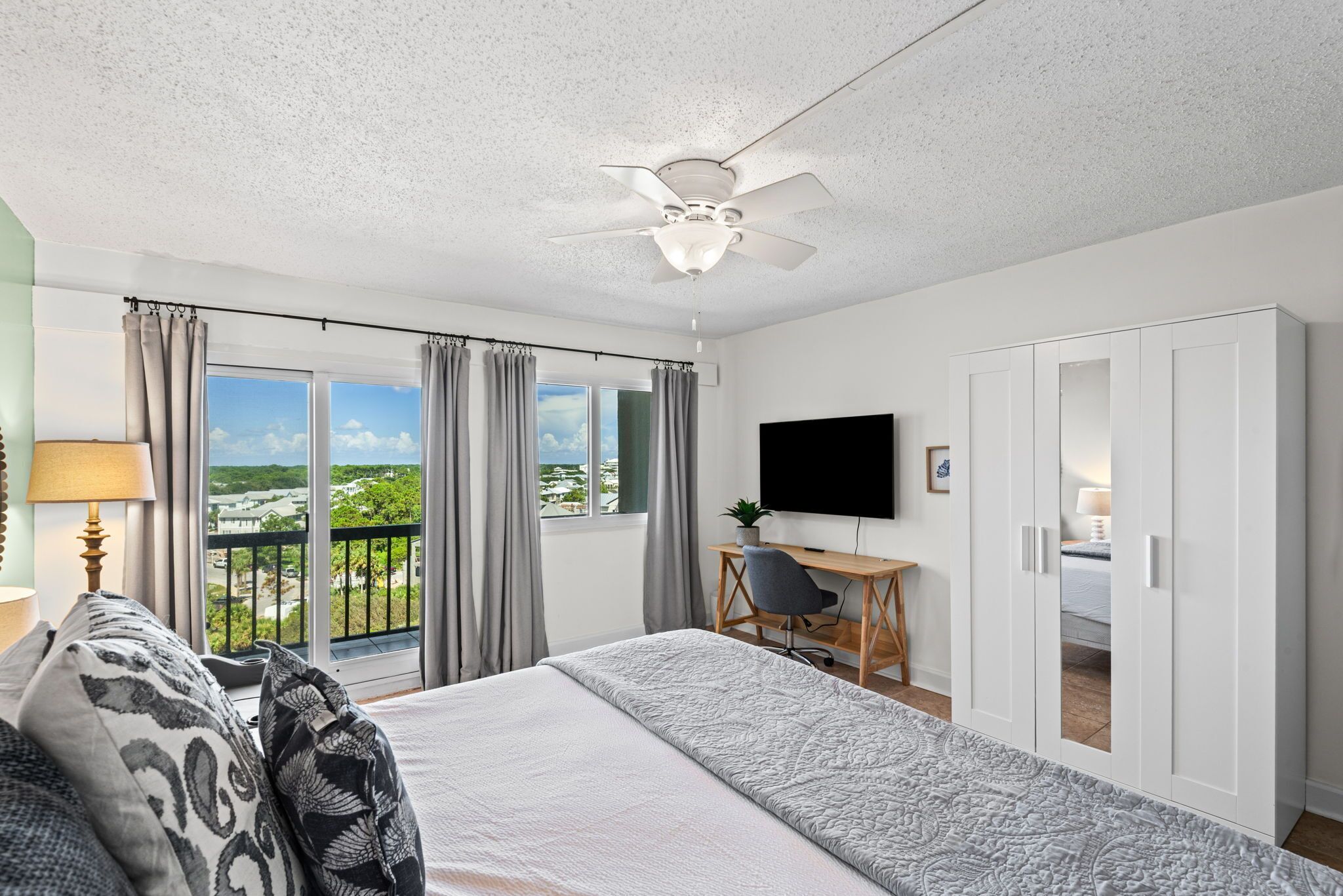 A bedroom with a bed, a desk, a chair, a TV, and a balcony with curtains.