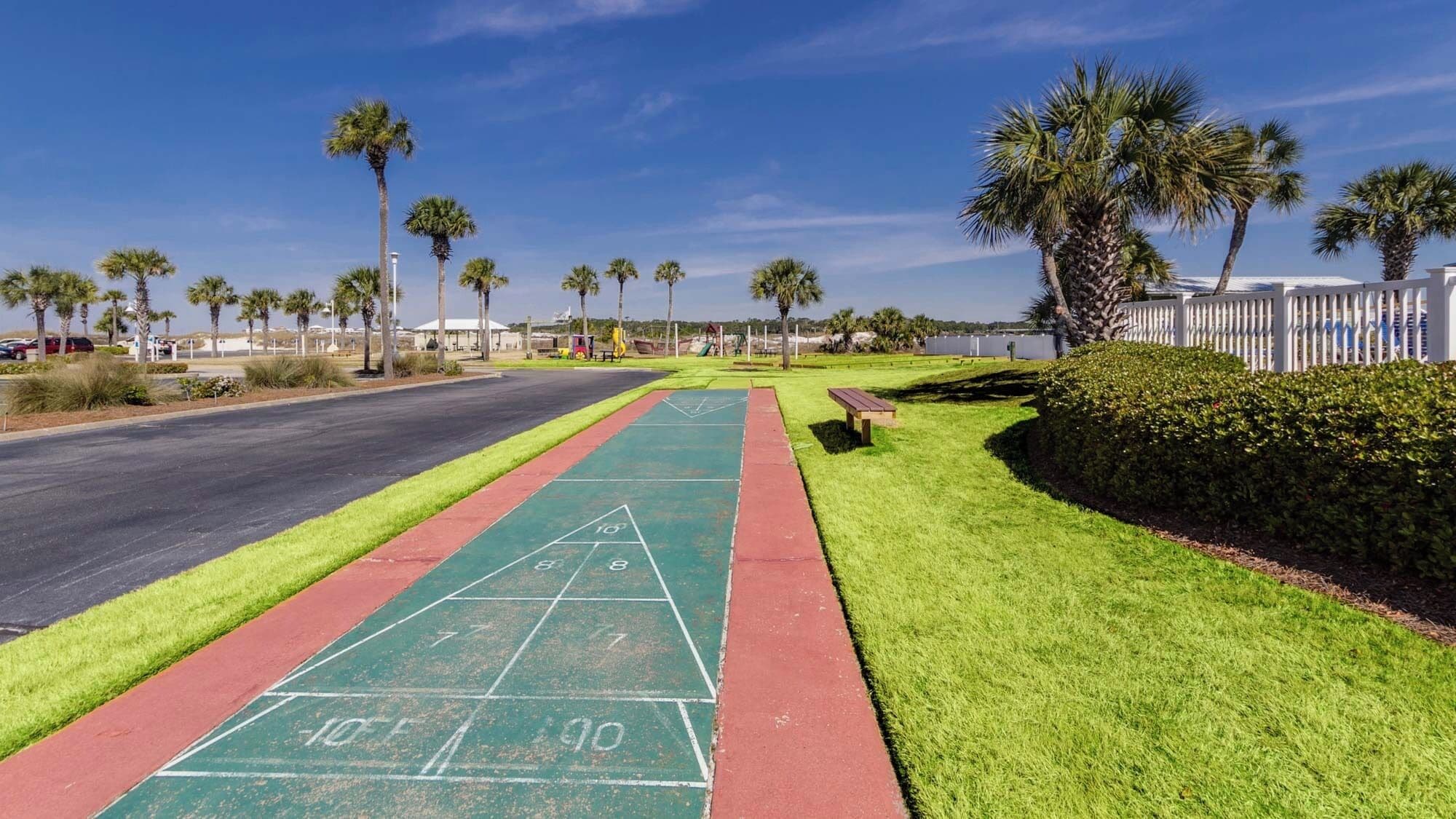 Ein sonniger Außenbereich mit einem grünen Shuffleboard-Spielfeld, einer roten Laufbahn und einem Parkplatz mit Palmen und einem weißen Zaun.