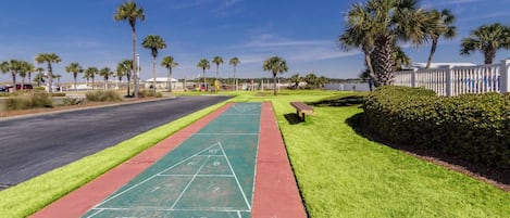 Ein sonniger Außenbereich mit einem grünen Shuffleboard-Spielfeld, einer roten Laufbahn und einem Parkplatz mit Palmen und einem weißen Zaun.