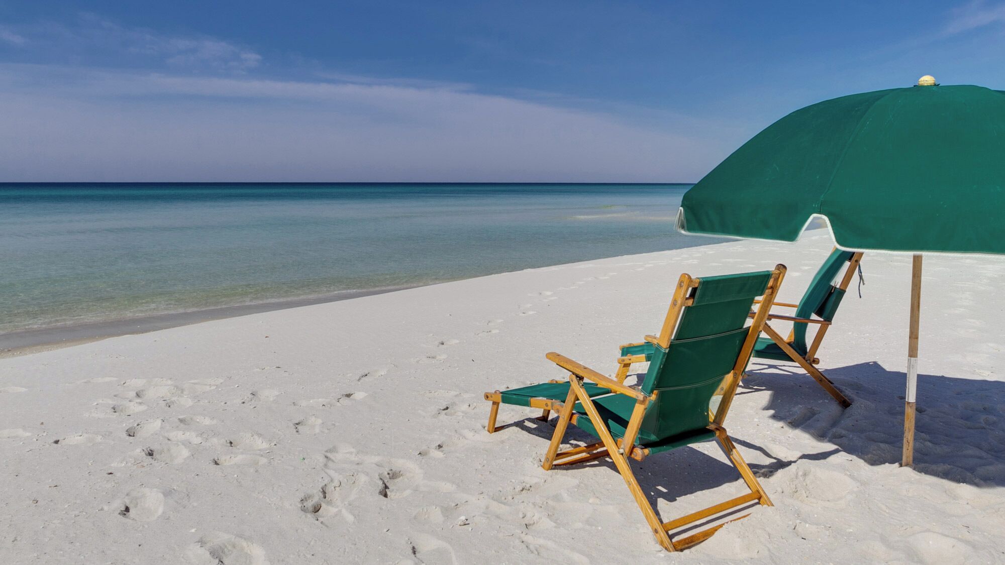 Una playa con dos sillas de madera y una sombrilla verde.