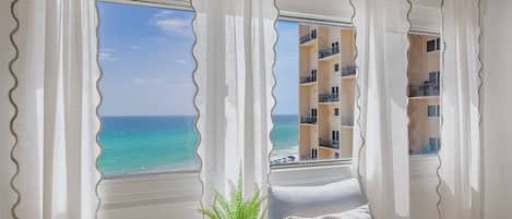 A room with a view of the ocean, a chair, a plant, and a window with curtains.