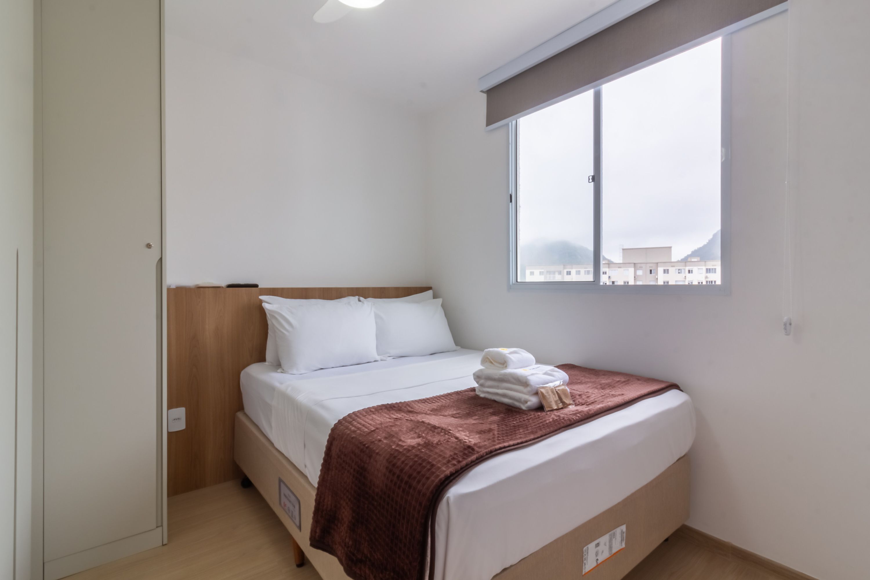 A single-bed room with a wooden headboard, white bedding, a brown throw blanket, and a window with a view of buildings.