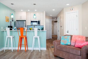Eine moderne Küche mit weißen Schränken, einem Edelstahlkühlschrank und einer Mikrowelle. Zwei Barhocker, einer orange und einer weiß. Im Wohnbereich steht ein braunes Sofa mit bunten Kissen.