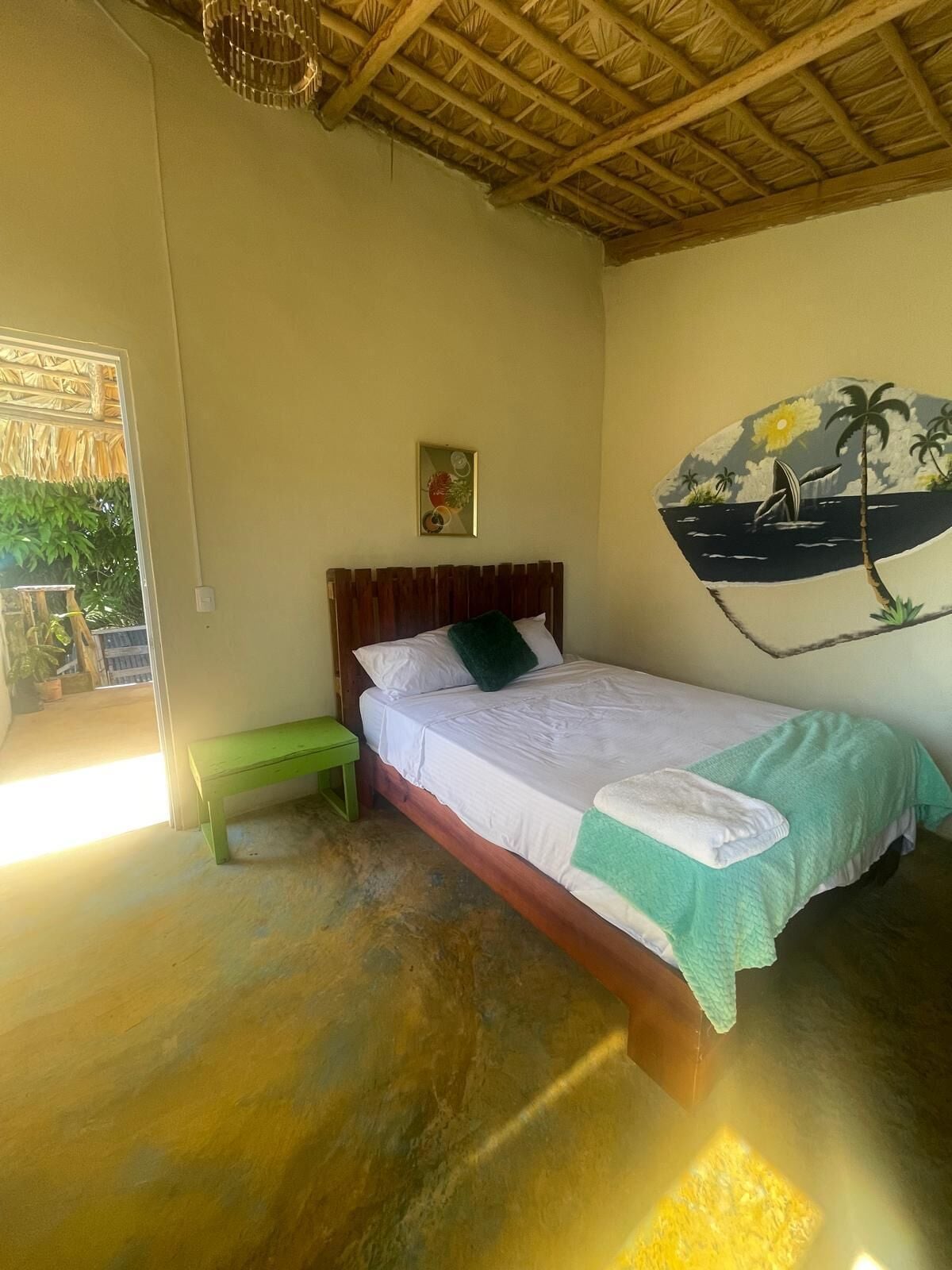 A room with a bed, a green bench, a wall with a tropical mural, and a thatched ceiling.