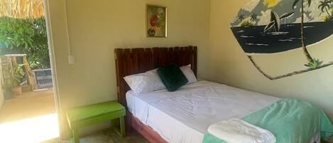 Una habitación con cama, un banco verde, una pared con un mural tropical y un techo de paja.