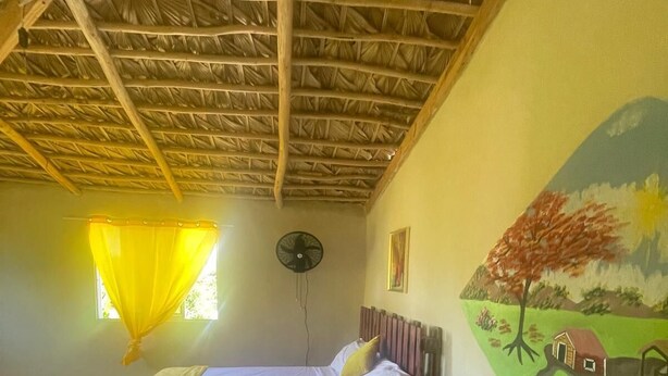 Ein Zimmer mit einem Bett, einem Sessel, einem Ventilator und einem Wandgemälde.