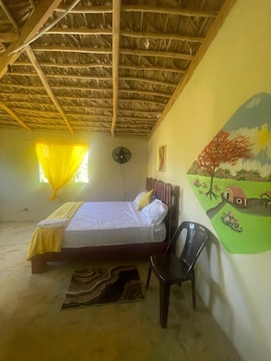 Una habitación con una cama, una silla, un ventilador y un mural en la pared.
