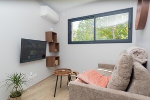A living room with a sofa, a TV mounted on the wall, a window with a view of trees, and a small table with a plant. - Les Nids des Mûriers (2-person) (CHAPAREILLAN)