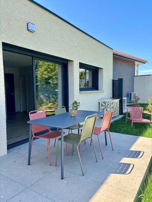 Une terrasse extérieure avec une table et des chaises, une porte-fenêtre coulissante donnant sur un espace abrité et un petit jardin avec des plantes.