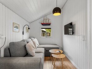 Un espacio habitable compacto con un sofá, una pequeña mesa redonda, un televisor montado en la pared y un modelo de barco colgando del techo.