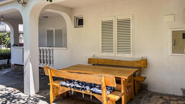 Un conjunto de mesa y bancos de madera frente a una pared blanca con contraventanas, bajo un porche cubierto.
