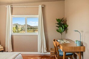 Ein Zimmer mit einem Bett, einem Holztisch mit Stuhl, einem Fenster mit Vorhängen, einer Zimmerpflanze und einer Lampe.