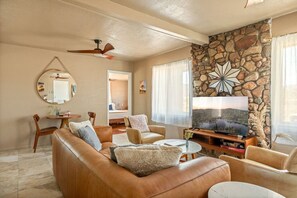 A living room with a stone accent wall, a large flat-screen TV, leather sofas, and a ceiling fan. - Aster Abode W/ Hot Tub & Game Room - 5 Mi to Park (Joshua Tree)