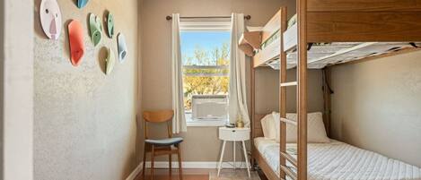 A compact room with a bunk bed, a chair, a small table, and a window with a view of trees.