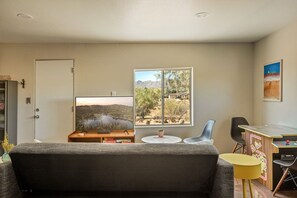 A living room with a sofa, TV, and a desk with a computer. There is a window with a view of a landscape. - Aster Abode W/ Hot Tub & Game Room - 5 Mi to Park (Joshua Tree)