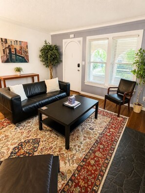 A living room with a black leather sofa, a coffee table, a patterned rug, and a door with a plant beside it. - Archer House 2BR · Workspace · Pets · Downtown (Tulsa)