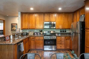 A kitchen with wooden cabinets, stainless steel appliances, granite countertops, and a bar area with chairs. - Pups and Pine Lodge | w/ BBQ Deck, Hot tub & Games (Big Bear)