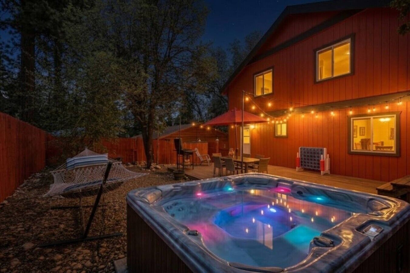 Un bain à remous avec des jeux de lumière colorés, une terrasse en bois et une maison décorée de guirlandes lumineuses.