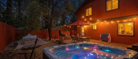 Un bain à remous avec des jeux de lumière colorés, une terrasse en bois et une maison décorée de guirlandes lumineuses.