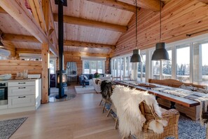 A log cabin interior with a central fireplace, dining area, and large windows.