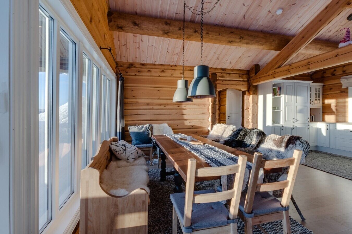 Interno di una baita in legno con zona pranzo, mobili in legno e cucina con pensili bianchi.