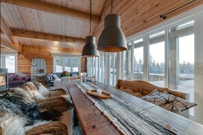 A spacious cabin interior with a long wooden dining table, multiple seating areas, and large windows offering views of a snowy landscape.