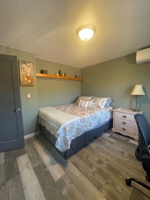 Een slaapkamer met een bed, nachtkastje, stoel, plank met decoratieve objecten en een deur.