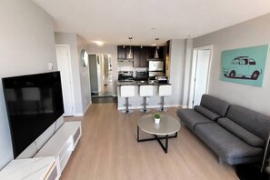 A modern living room with a flat-screen TV, a grey sofa, a small round table with a plant, and a kitchen in the background. - Panoramic False Creek View 2BR/2BA | Pool, Parking (Vancouver)