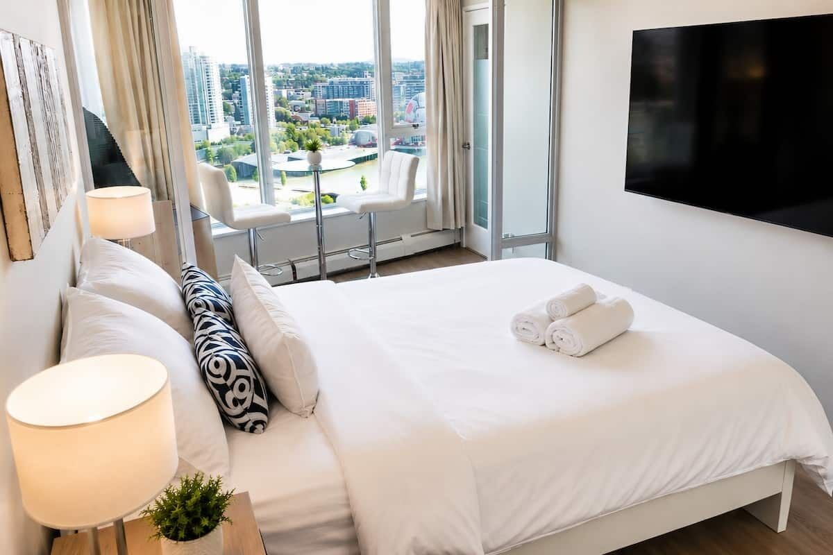 A hotel room with a bed, a TV, a window with a city view, and a chair by the window.