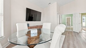 Ein modernes Wohnzimmer mit einem Esstisch mit Glasplatte, weißen Stühlen, einem flachbildfernseher an der Wand und einem Holzschrank.
