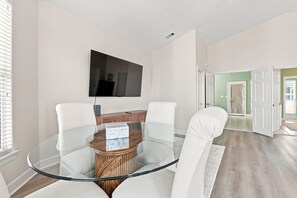 A modern living room with a glass-top dining table, white chairs, a flat-screen TV mounted on the wall, and a wooden cabinet. - 5BR 30A Beach House Private Beach Access - Sleeps 10 (Santa Rosa Beach)
