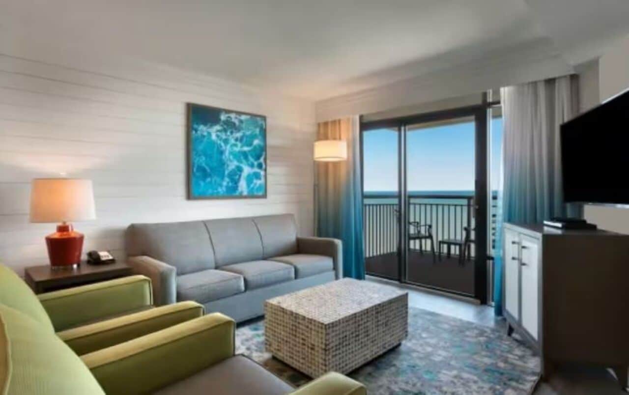 A hotel room with a sofa, a small table, a television, and a balcony with a view.