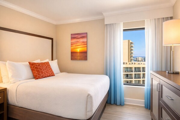 A hotel room with a bed, a nightstand, a painting of a sunset, a window with curtains, and a view of buildings. - Spacious Family Beach Stay | Pools, Hot Tubs & Ocean Views | Close to SkyWheel (Myrtle Beach)