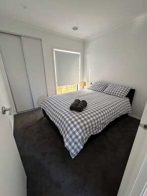 A bedroom with a bed, a window with blinds, a lamp, and a door. - Modern cozy home opp park in quiet neighbourhood (Cranbourne East)