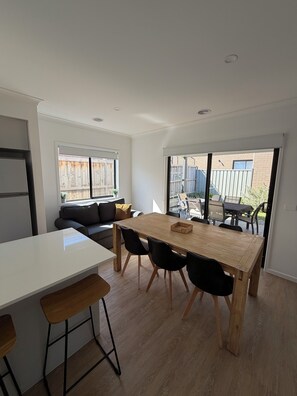 A modern kitchen with a central island, a dining area with a wooden table and chairs, and a view of the backyard through large windows. - Modern cozy home opp park in quiet neighbourhood (Cranbourne East)