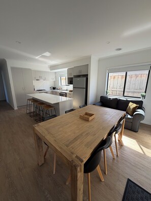 A modern kitchen with white cabinets, a central island, and a dining area with a wooden table and chairs. - Modern cozy home opp park in quiet neighbourhood (Cranbourne East)