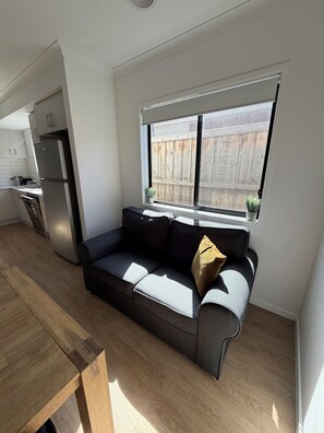 A compact living space with a kitchenette, a sofa with cushions, and a large window. - Modern cozy home opp park in quiet neighbourhood (Cranbourne East)