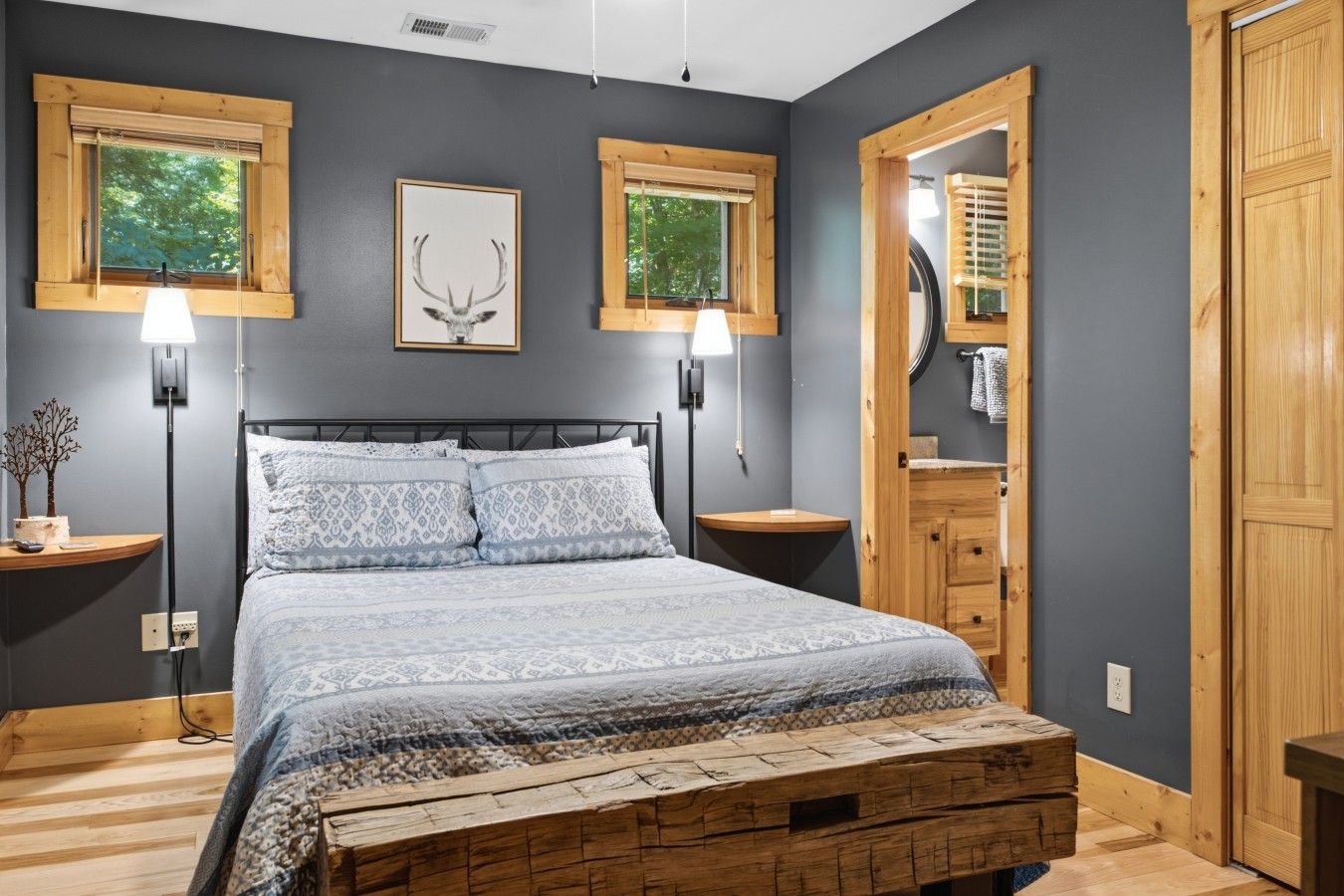 A bedroom with a bed, two bedside tables, a nightstand, a wooden bench, a wall-mounted lamp, and a framed deer head picture.