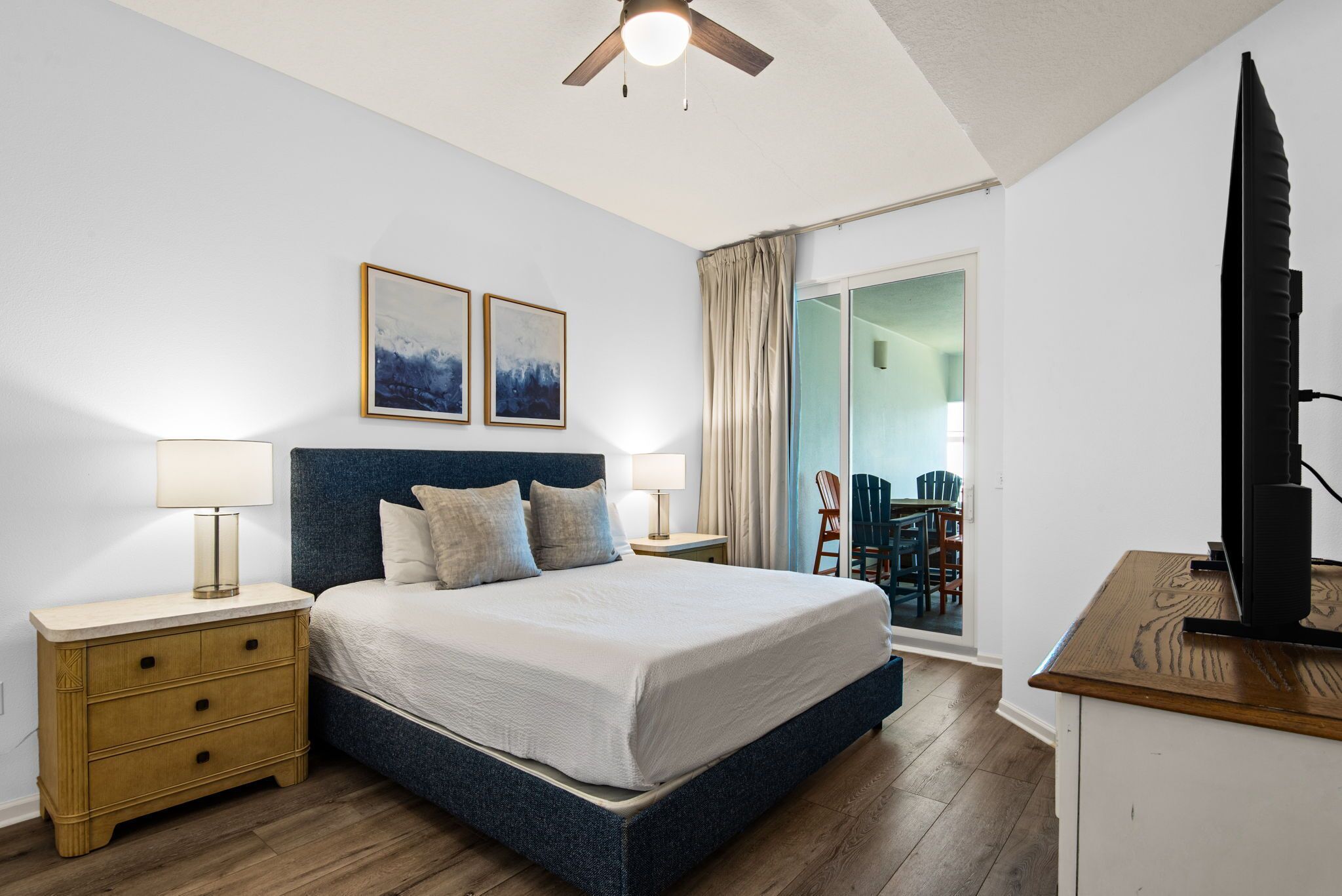 A bedroom with a bed, bedside tables, a ceiling fan, and a view of an outdoor area with chairs.
