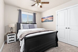 A bedroom with a bed, a nightstand, a lamp, a ceiling fan, a window with curtains, a door, and a wall-mounted picture.