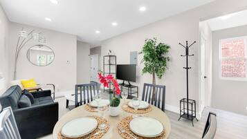 A modern living room with a round dining table set for four, a sofa, a TV, and a coat rack.