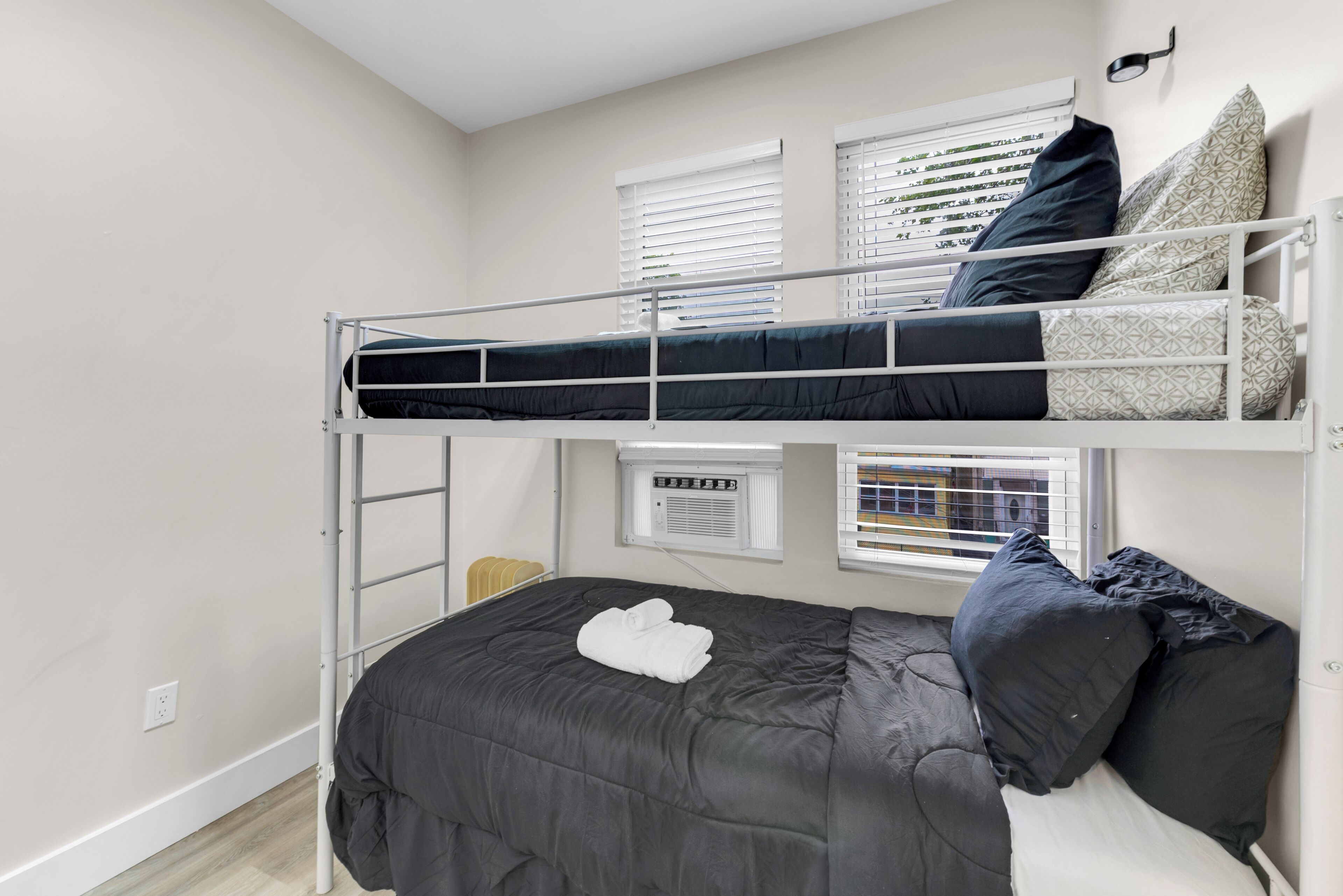 A bunk bed with a lower bed and a ladder, a window with blinds, and a wall-mounted air conditioning unit.