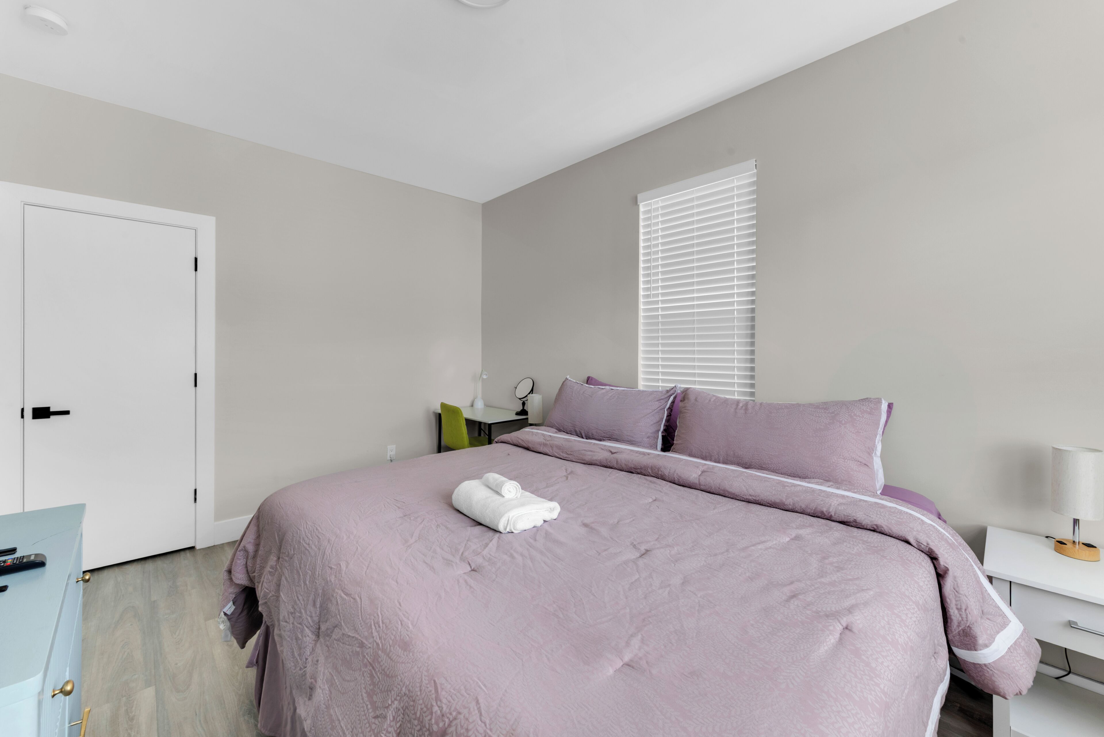 A bedroom with a bed, a desk, a chair, a window with blinds, and a door.