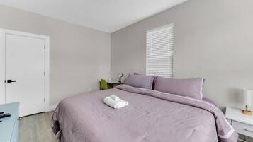 A bedroom with a bed, a desk, a chair, a window with blinds, and a door.